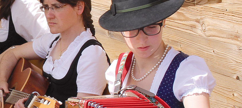 Zwei Frauen in Tracht vor einer Hütte musizieren an Gitarre und Ziehharmonika