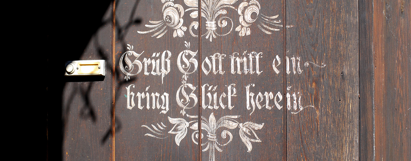 eine Holztür auf welcher in weißer Schrift "Grüß Gott tritt ein bring Glück herein" steht. 